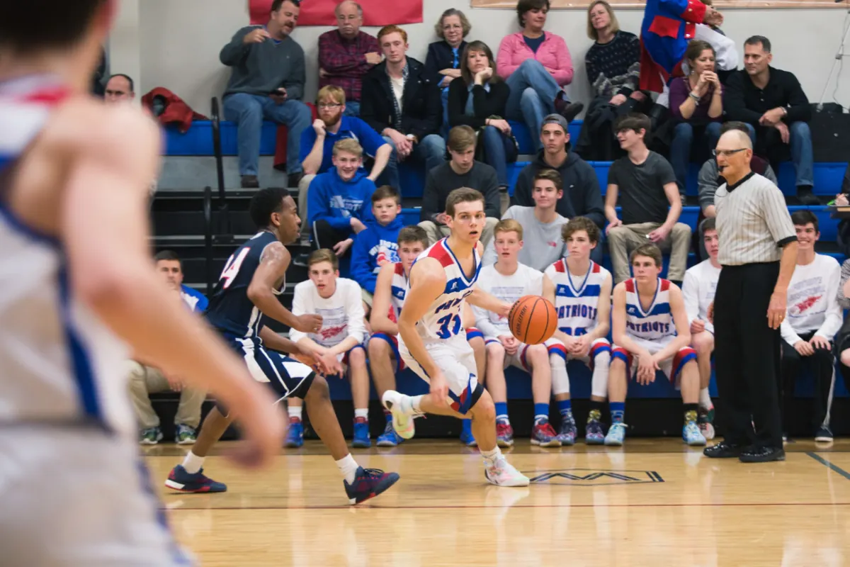 A Patriots basketball player driving up the court in front of the bench and a packed home crowd