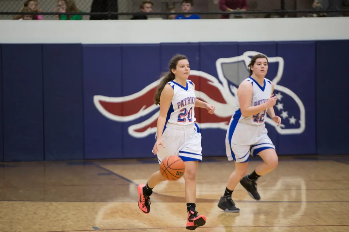 Two Patriots girls basketball players bringing the ball up the court in front of the Patriots logo
