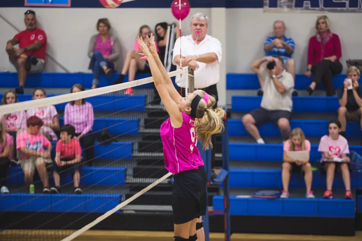 A Patriots volleyball player going up for a block at the net in front of a pink-out crowd