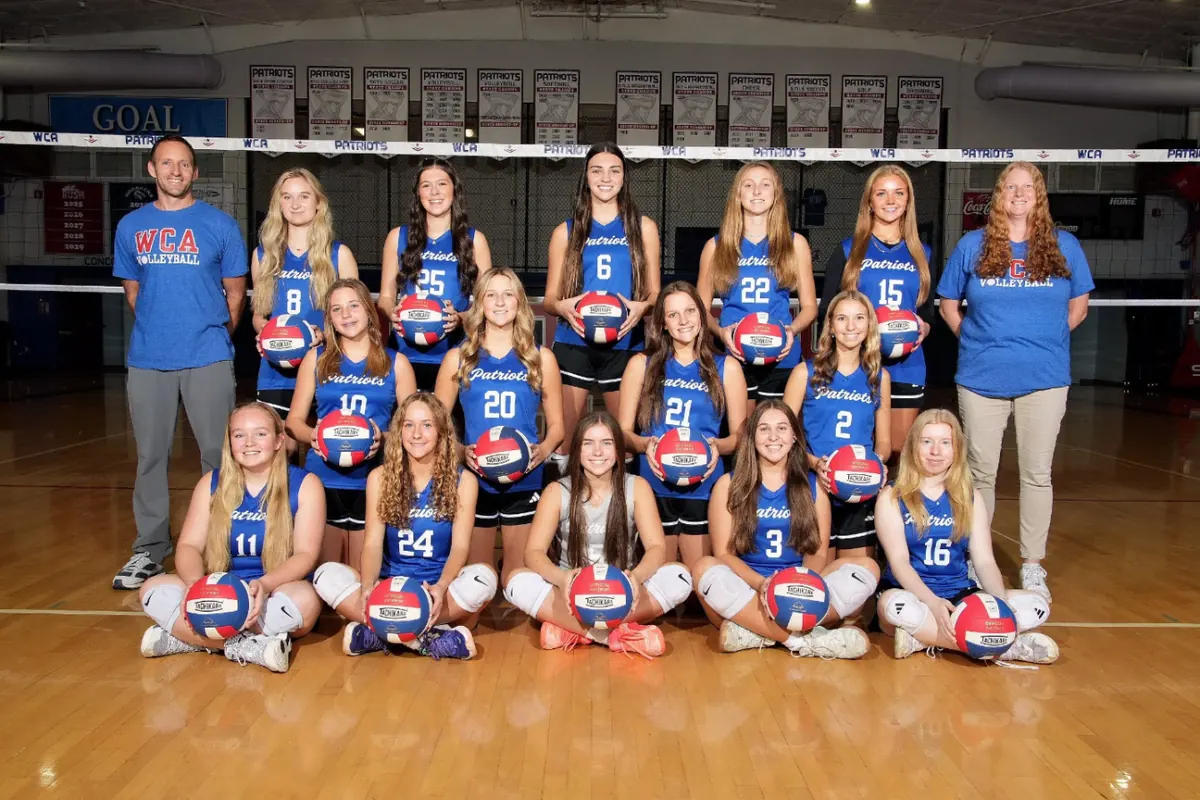 The varsity volleyball team and coaches in the gym under the Patriots state championship banners