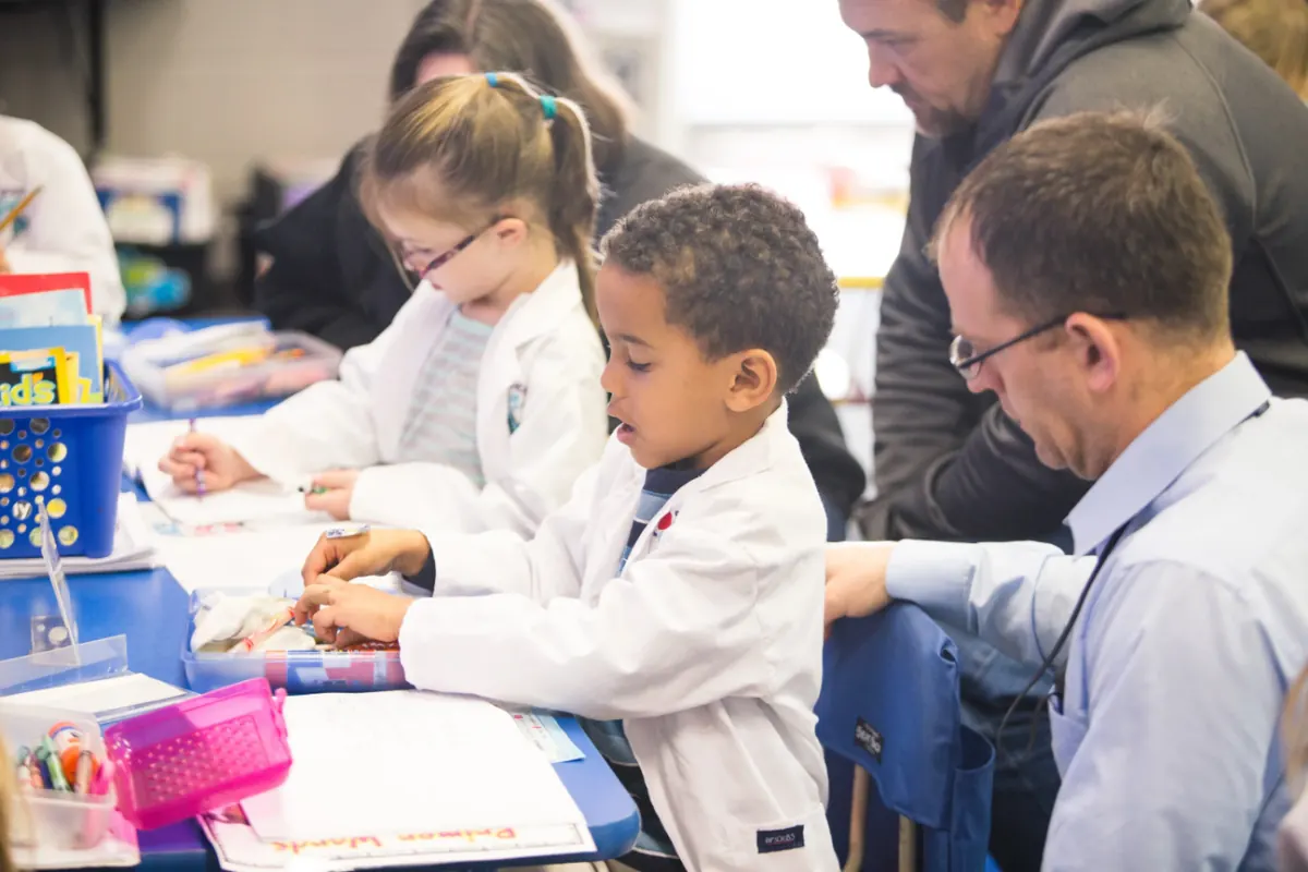 Kindergartners in lab coats working on a science activity as parents look on
