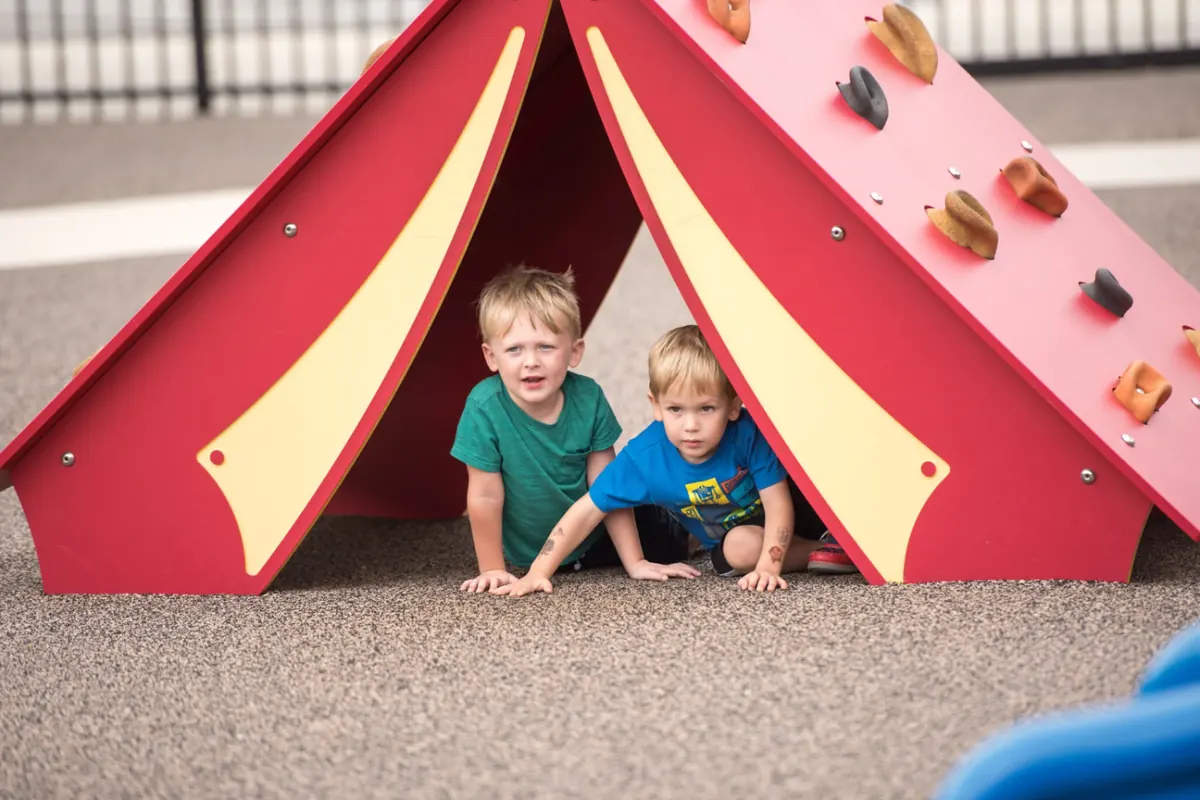 Two preschool boys peeking out of a red play tent on the WCA playground