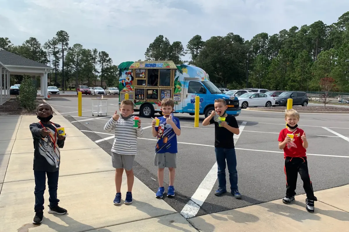Five Summer Day Camp boys giving a thumbs up with cups of shaved ice in front of a Kona Ice truck on campus