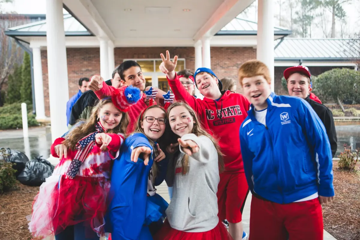 WCA students in red, white, and blue spirit wear cheering and pointing at the camera outside a campus building