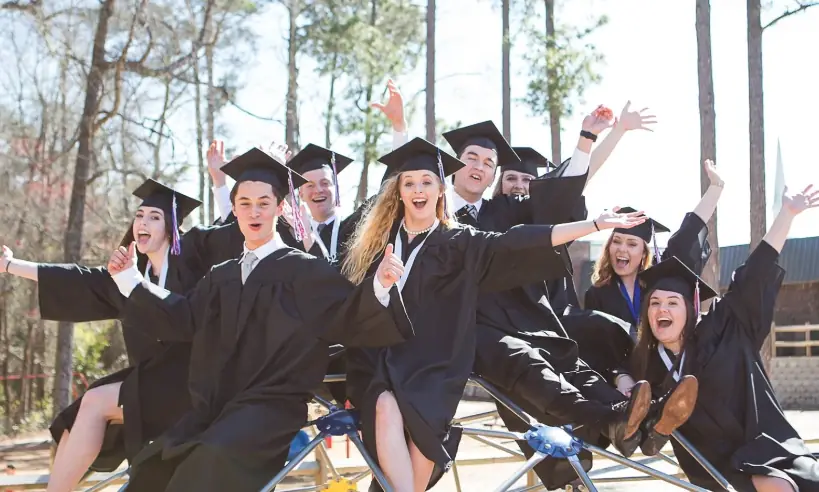 WCA graduates in caps and gowns celebrating on the campus playground, arms raised